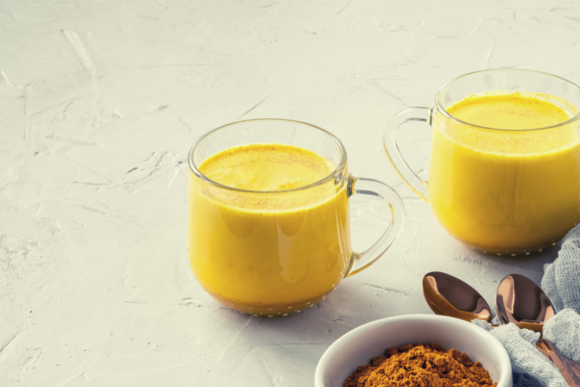 dwie-przezroczyste-szklanki-ze-zlotym-mlekiem-biala-miseczka-ze-zmielonym-cynamonem-metalowe-lyzki-z-cynamonem-na-jasnym-tle