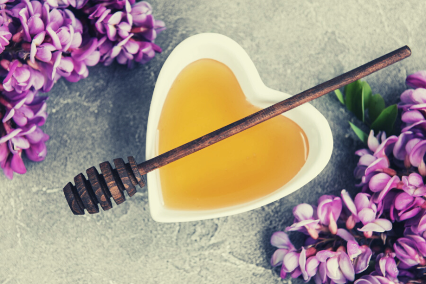 miod-i-lyzka-do-miodu-w-bialej-miseczce-w-ksztalcie-serca-na-szarym-tle-obok-fioletowe-kwiaty-akakcji