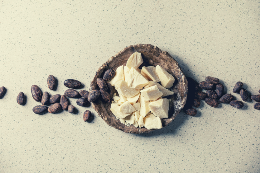 maslo-kakaowe-w-kosmetykach-poznaj-jego-wlasciwosci-maslo-kakaowe-na-brazowym-talerzu-ziarna-kakaowca-na-jasnoszarym-tle-widok-z-gory