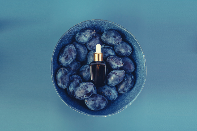 olej-z-pestek-sliwki-wielozadaniowosc-w-jednym-kosmetyku-niebieska-miska-z-dojrzalymi-sliwkami-na-nich-serum-w-butelce-z-ciemnego-szkla-z-pipetka-niebiesko-fioletowe-jednolite-tlo-widok-z-gory